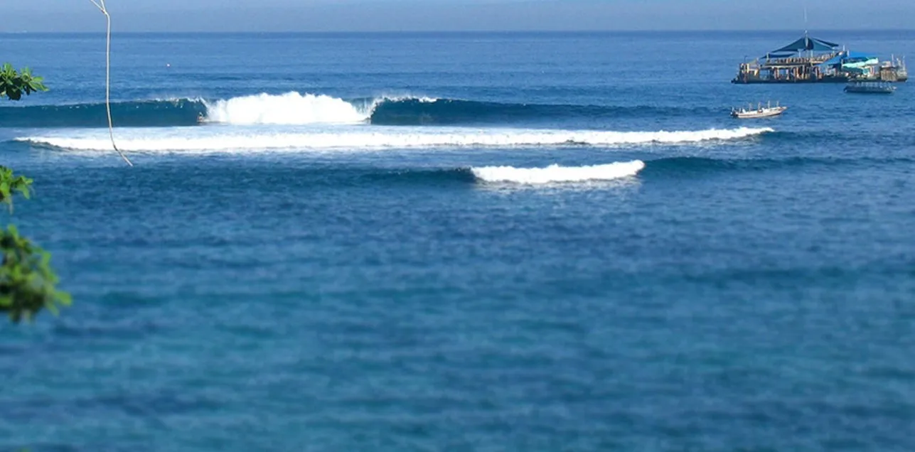 A clean wave peeling across deep blue water at Playgrounds, Nusa Lembongan, with a floating warung and a boat out in the channel